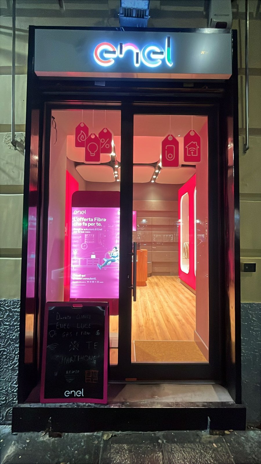 Vetrina notturna dello Spazio Enel Partner Napoli Corso Garibaldi 133: insegna Enel illuminata sopra la vetrina, doppia porta a vetri, illuminazione magenta interna del format Linea Unique di Enel Energia, totem digitale con offerta Fibra Enel Energia, lavagna promozionale sul marciapiede