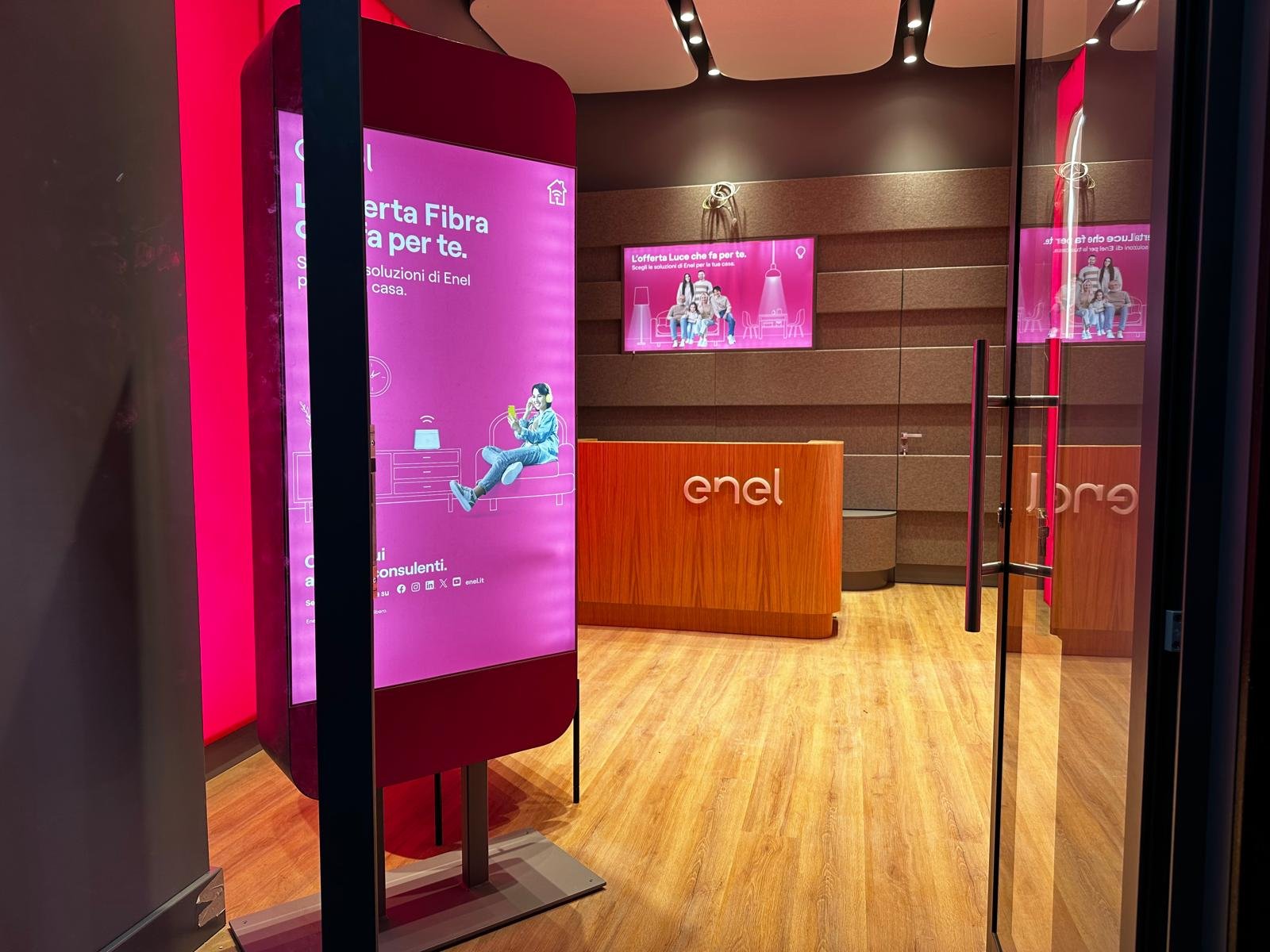 Interno dello Spazio Enel Partner Napoli Corso Giuseppe Garibaldi 133: totem digitale magenta con offerta Fibra Enel Energia, banco reception in legno con logo Enel, display digitali su parete con offerte Luce, pavimento in parquet chiaro