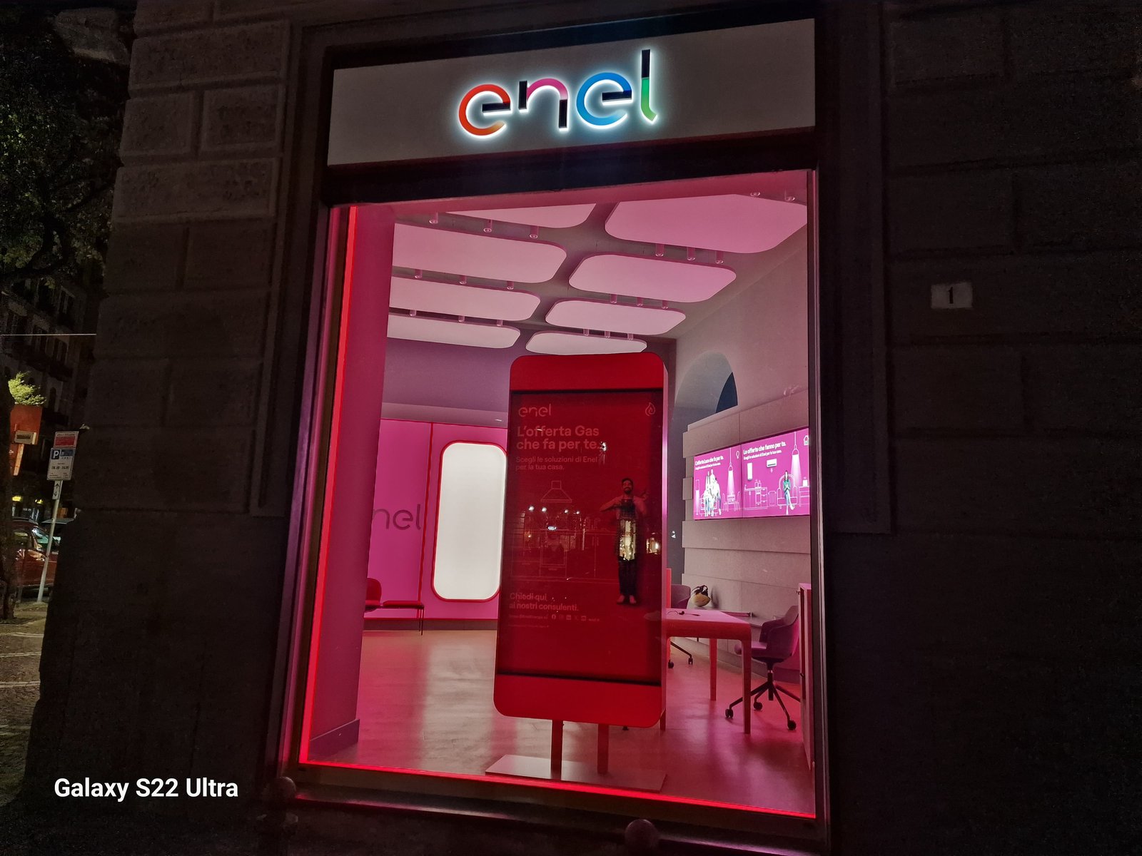 Vetrina notturna dello Spazio Enel Partner Napoli Corso Umberto I 123: insegna Enel illuminata sopra la vetrina fronte strada del Rettifilo, illuminazione magenta interna del format Linea Unique, totem digitale con offerta Gas Enel Energia