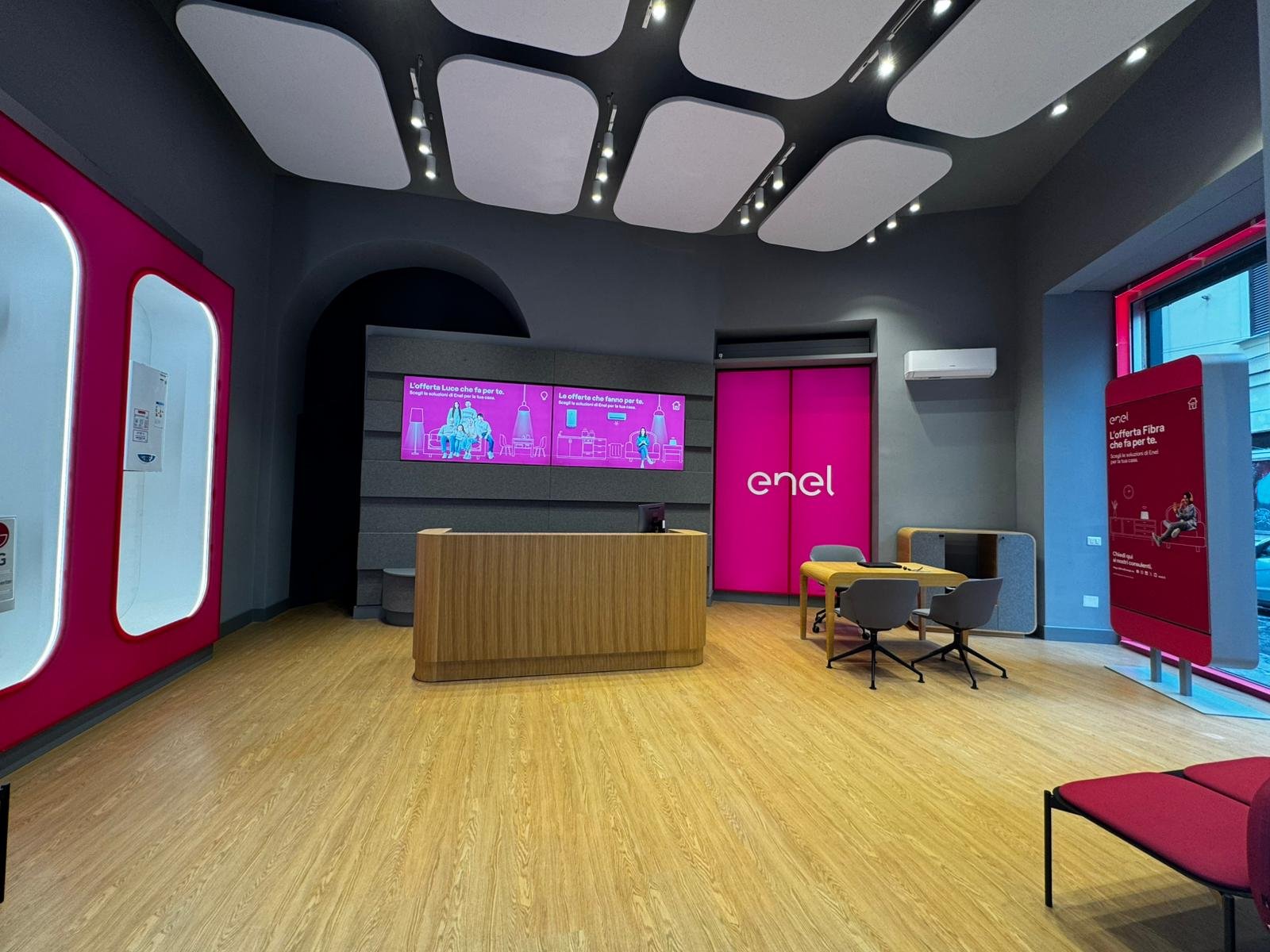 Interno dello Spazio Enel Partner Napoli Corso Umberto I 123: banco reception centrale in legno con monitor, area consulenza con tavolo e sedie, display digitali su parete con le offerte Luce Gas Fibra Enel Energia, soffitto Linea Unique con pannelli sagomati, pavimento in parquet, altezza 6 metri