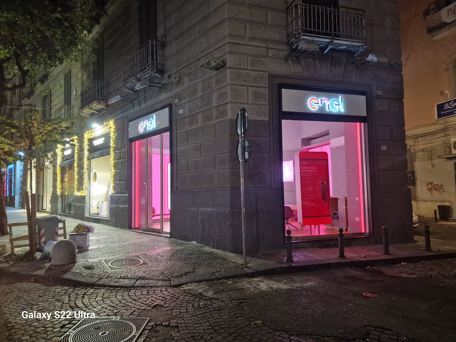 Vista d'angolo notturna dello Spazio Enel Partner Napoli Corso Umberto I 123: negozio all'angolo con doppia vetrina su due lati dell'edificio, entrambe con insegna Enel illuminata, illuminazione magenta interna, pavimentazione in cubetti di pietra del Rettifilo, cornice urbana tipica del centro storico di Napoli