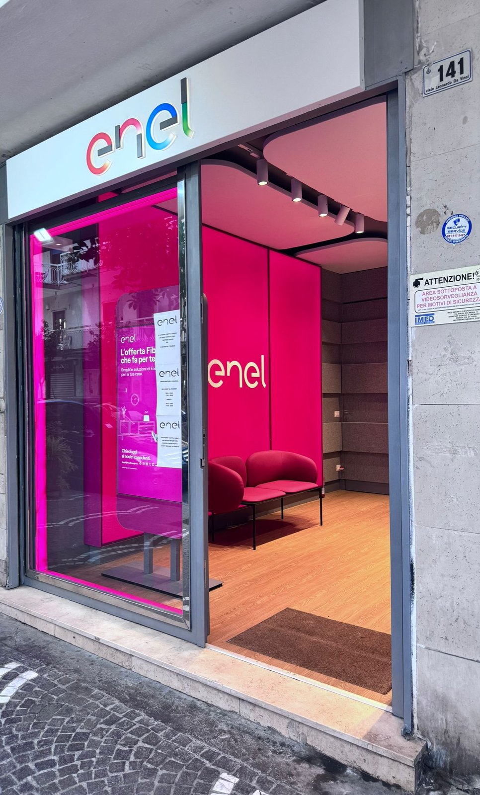 Interno dello Spazio Enel Partner Portici Viale Da Vinci 141: banco reception centrale in legno con logo Enel bianco luminoso, display TV a parete con offerta Luce Enel Energia, pareti magenta con curve arrotondate tipiche del format Linea Unique, espositore climatizzatori Comfee, pavimento in parquet chiaro