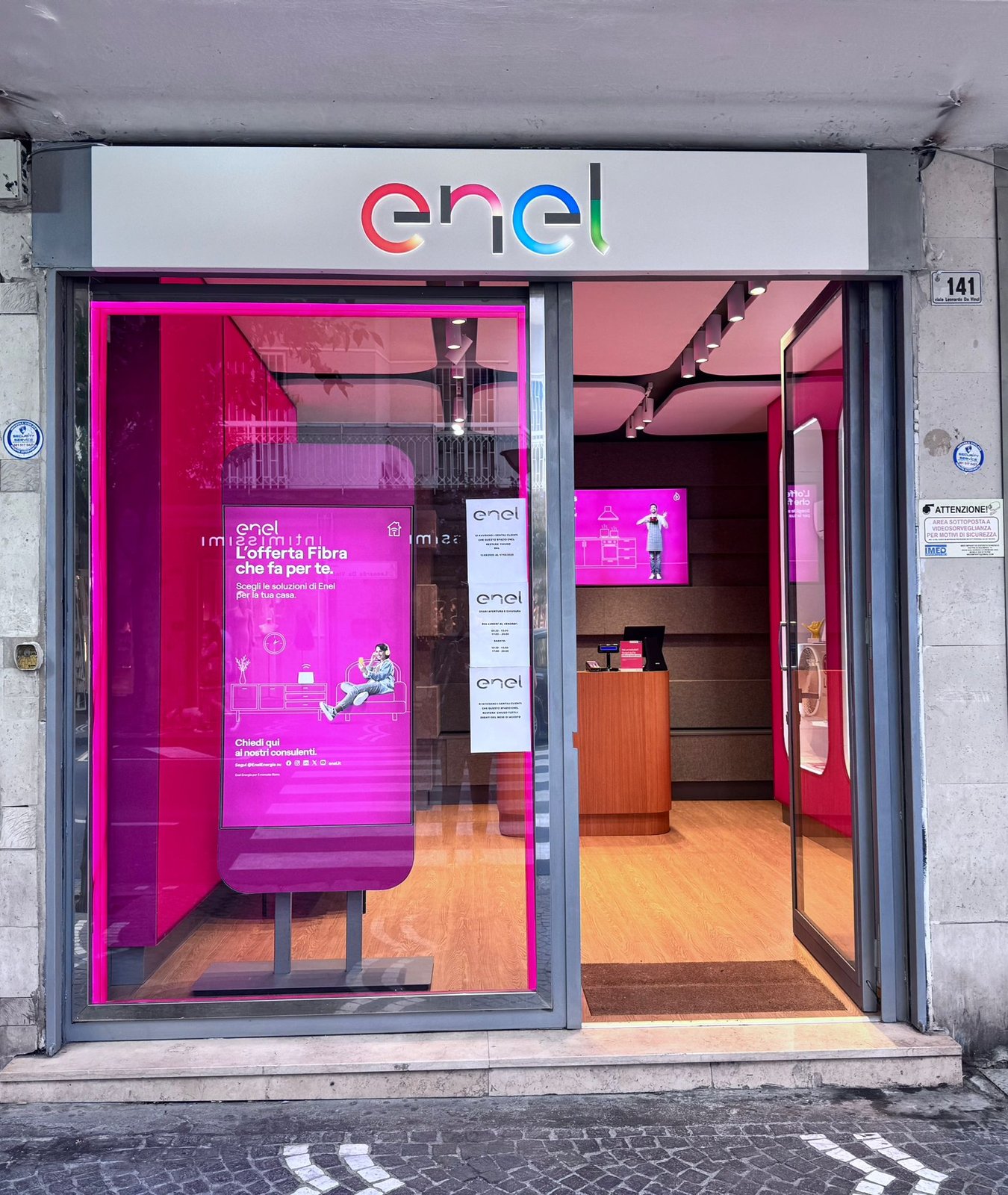 Facciata giorno dello Spazio Enel Partner Portici Viale Leonardo da Vinci 141: insegna Enel colorata sopra la vetrina, totem digitale con offerta Fibra Enel, ingresso con porta a vetri